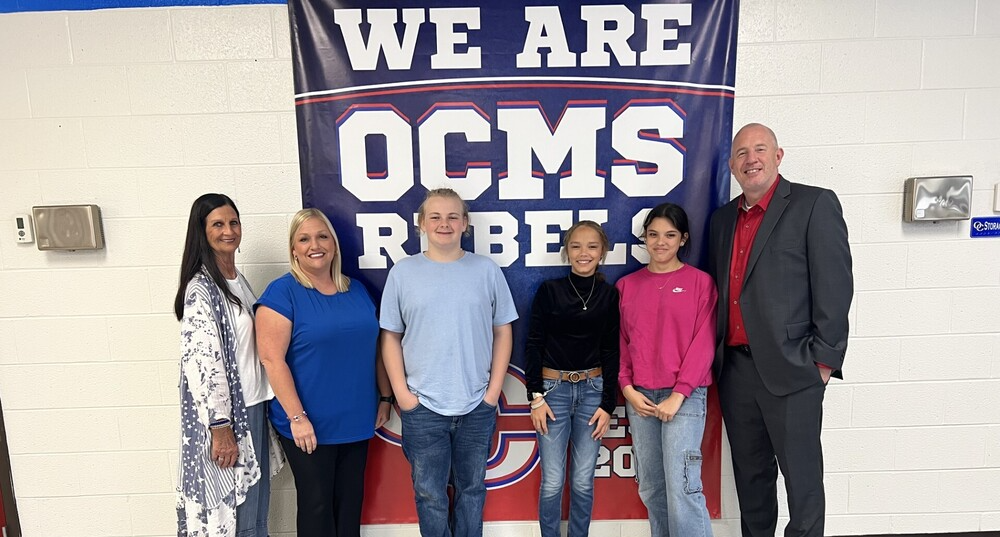 Six people pose together in front of an “OCMS Rebels” banner.