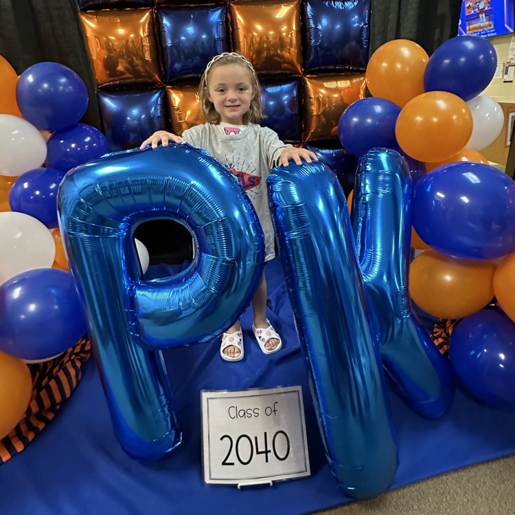 Young Student standing in front of a LRS backdrop holding “PK” balloon letters.
