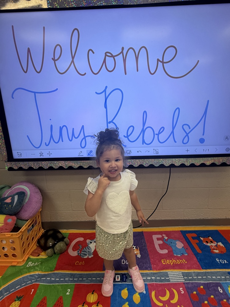 A young child standing in front of a smart board stating “Welcome Tiny Rebels!"