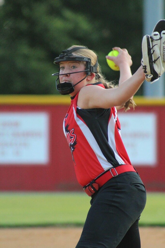 A female softball pitcher in a red, white, and black uniform throws a yellow softball toward home plate on a field, with a catcher’s glove visible and trees and a red outfield fence in the background.