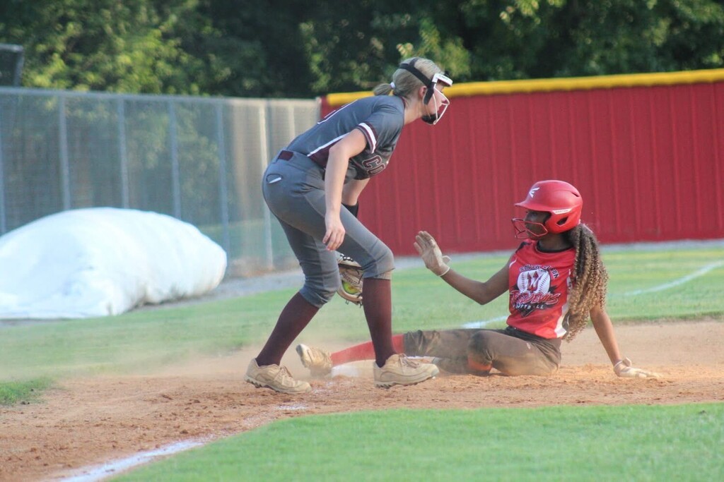 A softball player in a red uniform slides into a base as a fielder in gray reaches toward her, kicking up a cloud of dust.