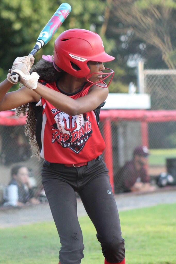 A softball player in a red helmet and Red Devils uniform holds a bat ready to swing during a game.