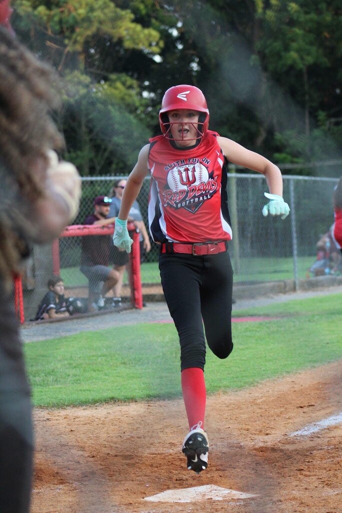 A softball player wearing a red helmet and “South Fulton Red Devils” uniform runs toward home plate during a game, with a catcher partially visible in the foreground.