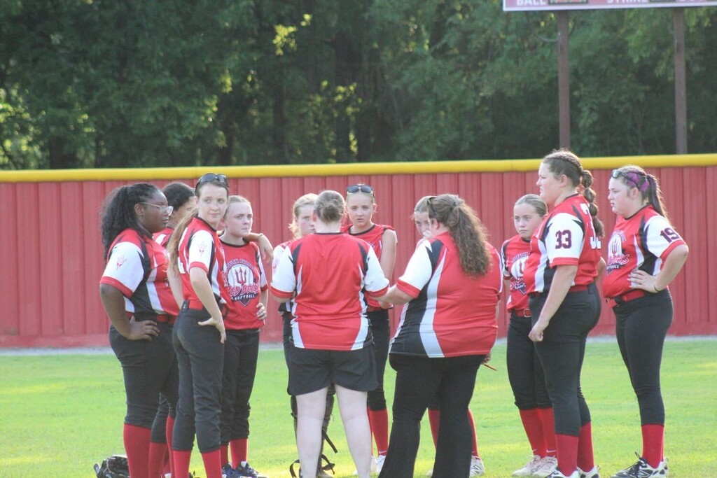 A girls’ softball team gathers in a huddle on the field, wearing matching red, white, and black uniforms, with a wooded area and red outfield fence in the background.