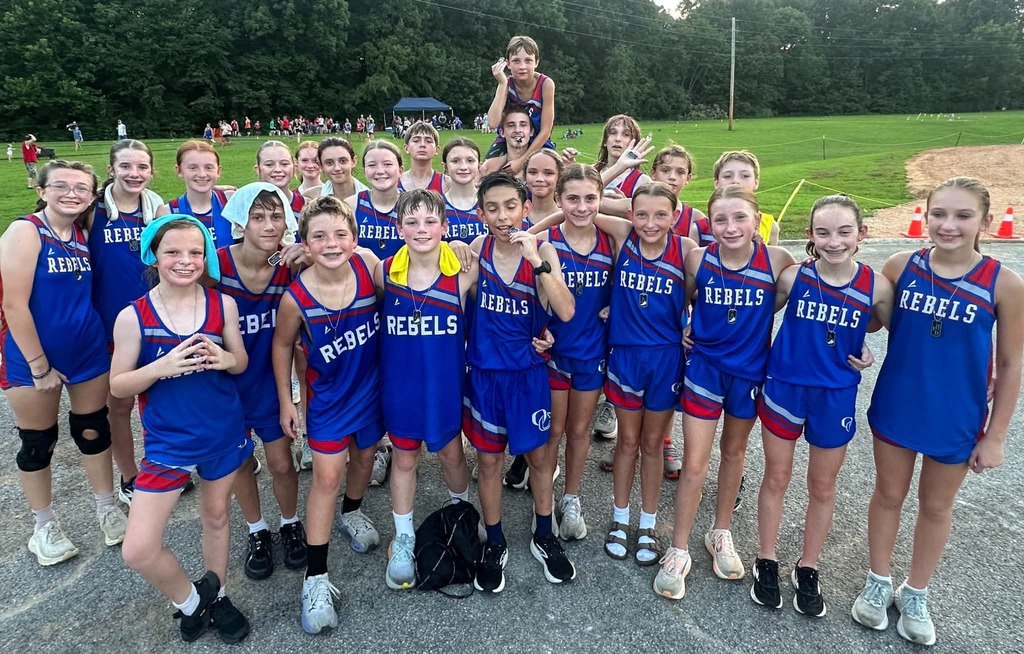 A large group of young athletes wearing matching blue-and-red “REBELS” uniforms pose together outdoors at a track-and-field event. They smile and stand closely together on a paved area, with a grassy field, trees, spectators, and athletic equipment visible in the background. One boy sits on another teammate’s shoulders near the center of the group.