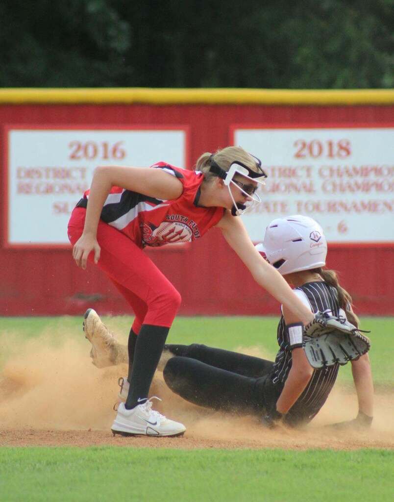 A softball player in red slides into home as a catcher in black reaches for the ball.