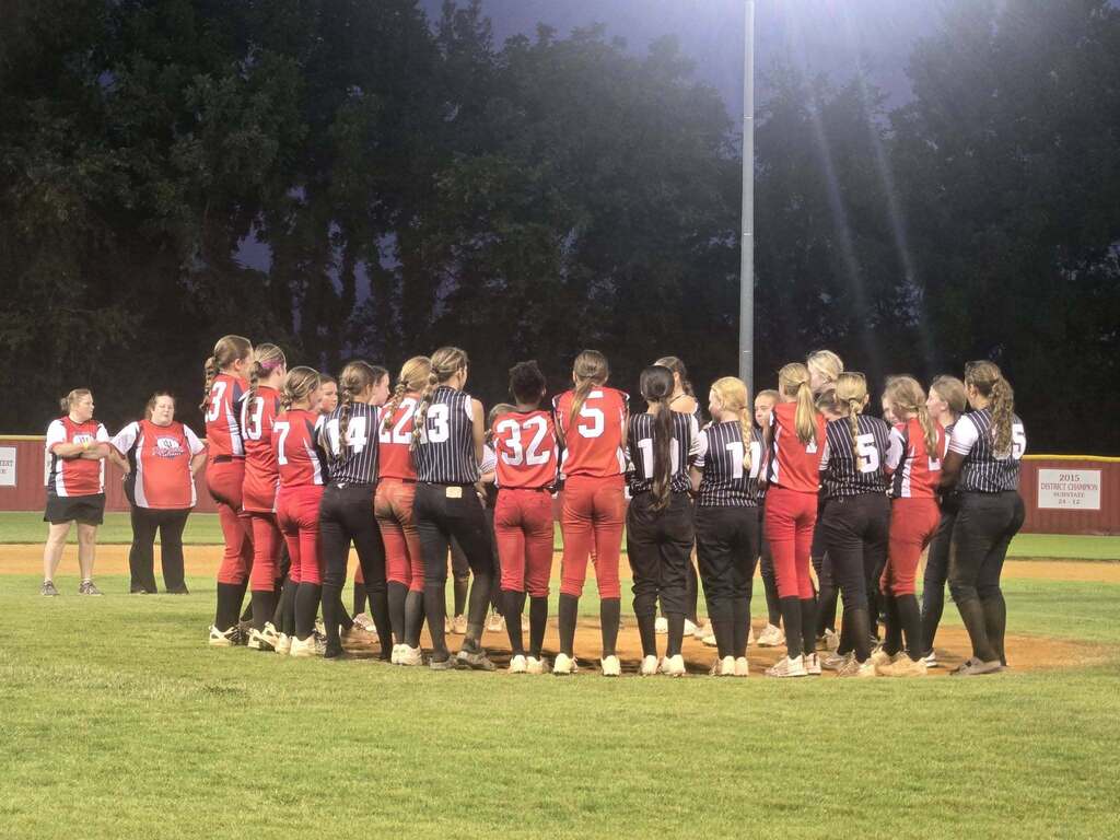 A group of girls in softball uniforms gathers on a baseball field at night.