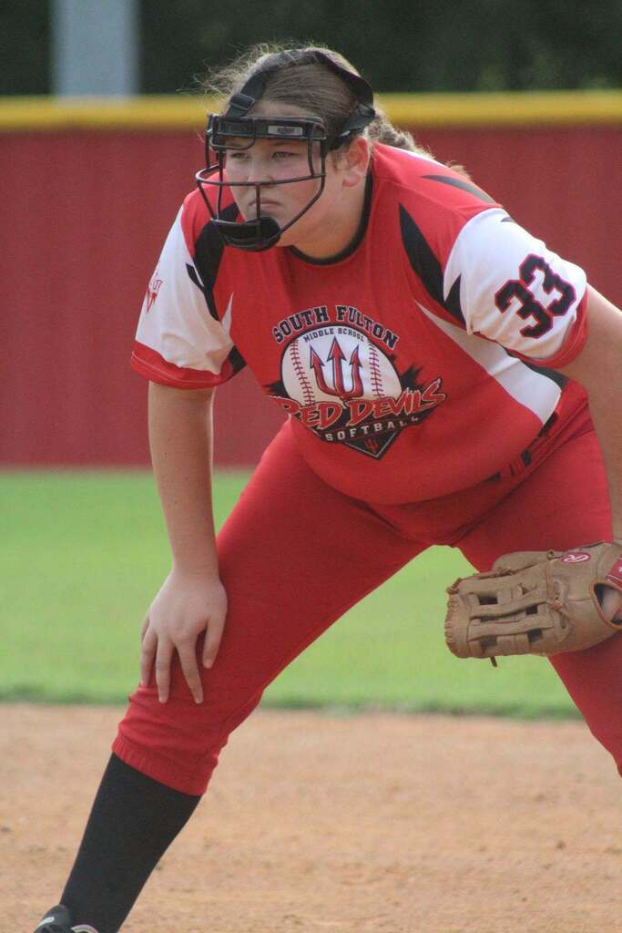 A softball player in a red uniform and helmet crouches in a ready position on the field.
