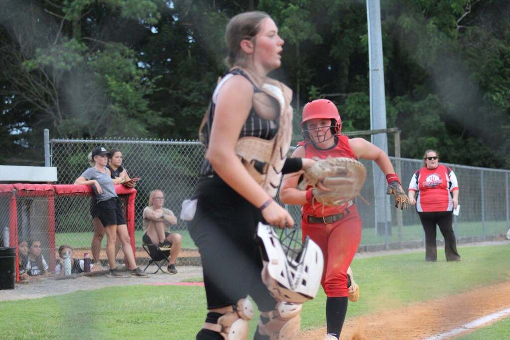 A softball catcher in red gear prepares to catch the ball as a batter runs toward home plate.