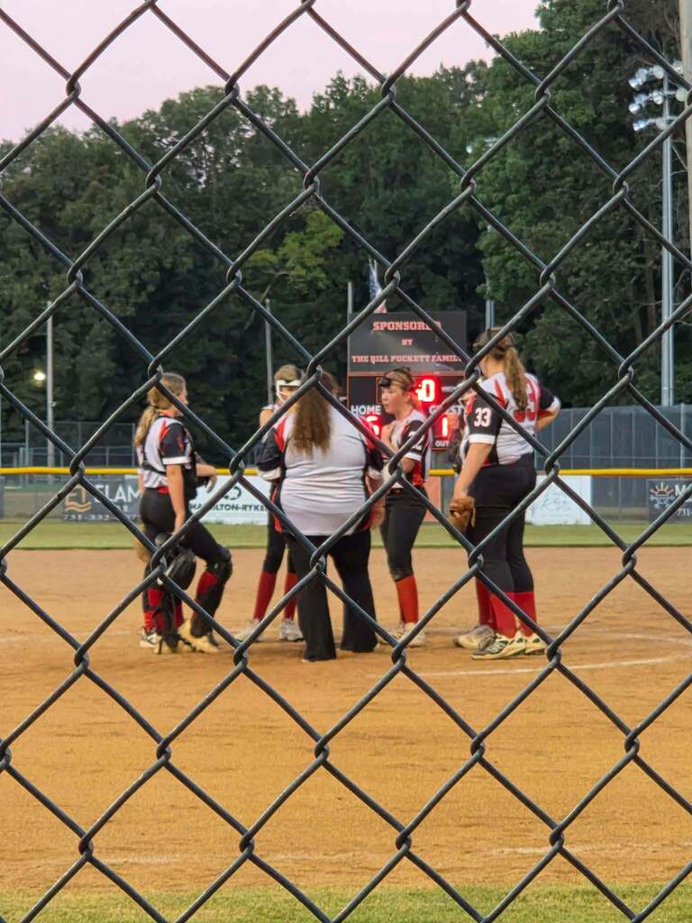 A group of softball players gathers near home plate during a game, viewed through a chain-link fence, with the scoreboard and field in the background.
