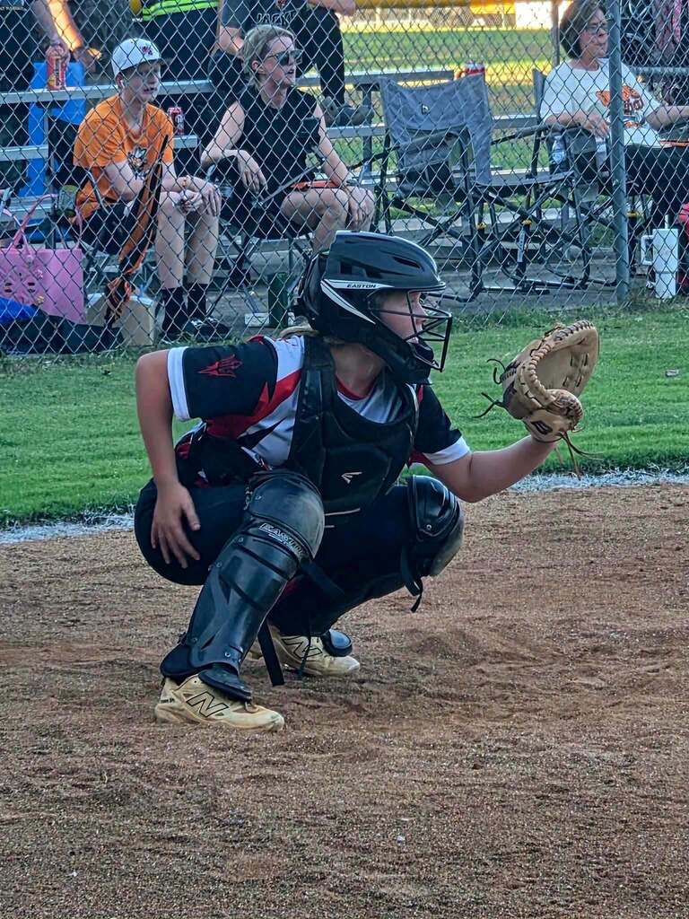 A young softball catcher crouches on the field wearing protective gear and holding a glove. Spectators sit behind a chain-link fence.