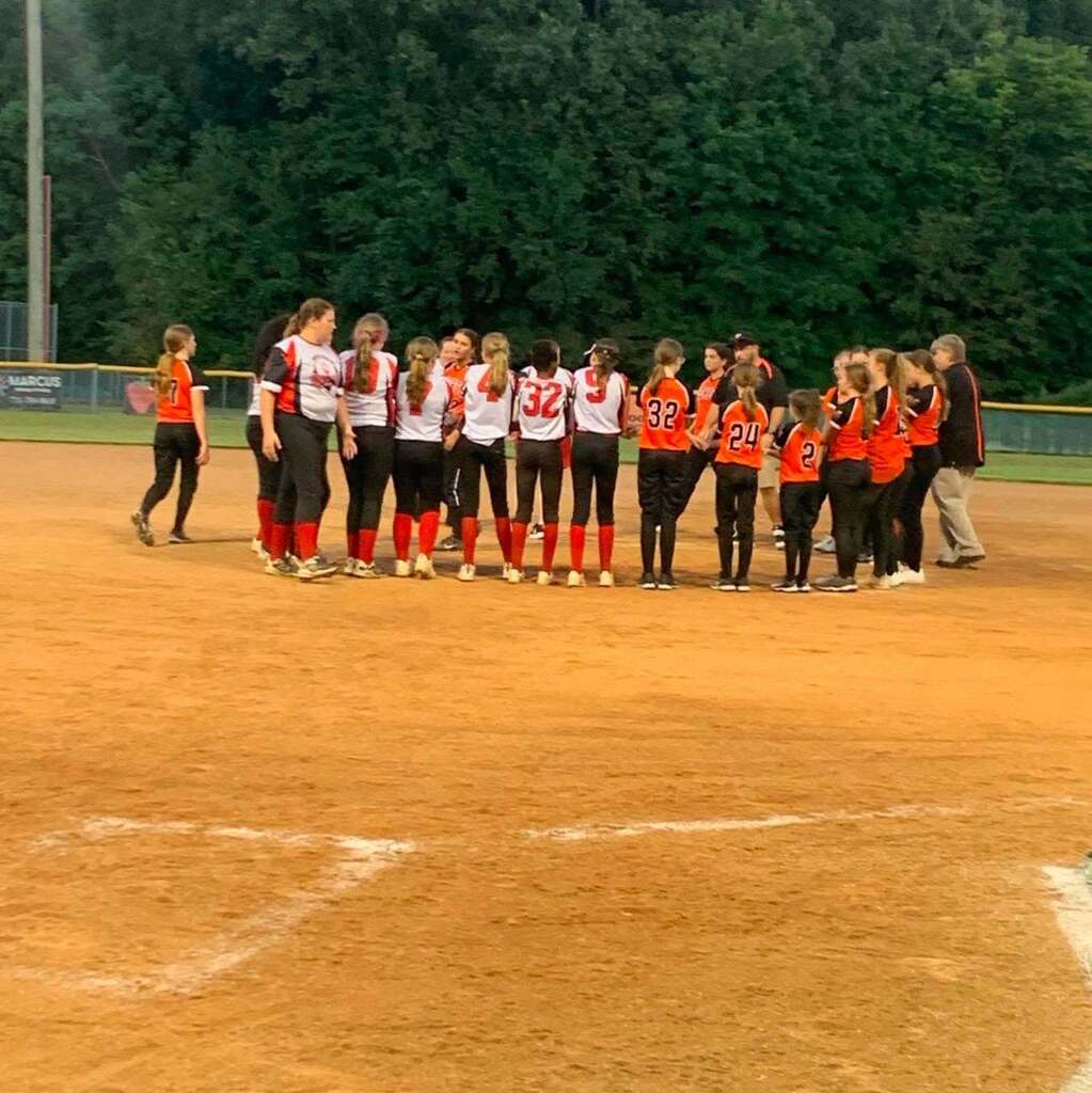 A group of girls in softball uniforms gathers on a dirt field during a game.
