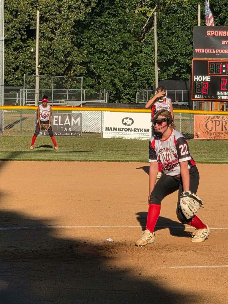 A softball player in uniform and protective gear stands on the infield during a game, with another player and the scoreboard visible in the background.