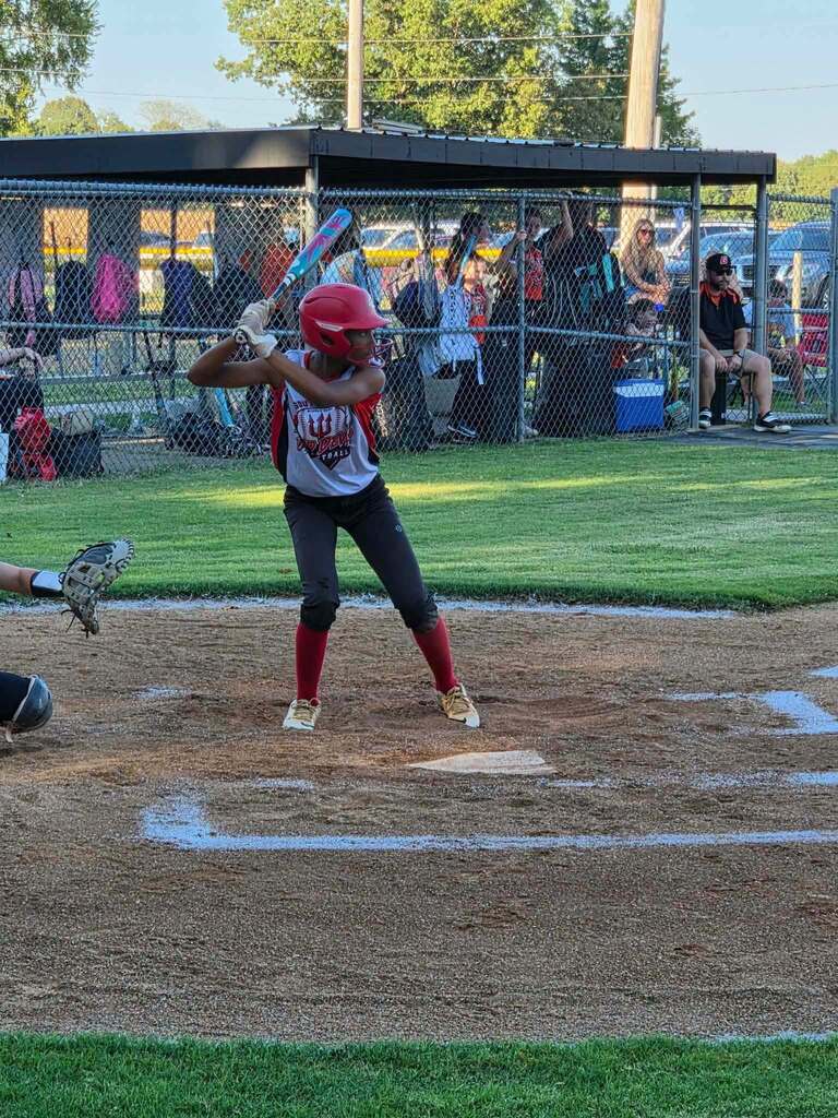 A young softball player in a red helmet and uniform prepares to swing at a pitch, with a catcher and spectators behind the fence.