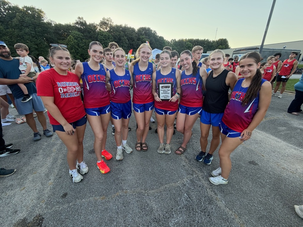 OCCHS girls cross country team poses with their first-place team plaque following the Dresden Invitational. Several runners stand together in blue and red uniforms, with a supporter wearing a “Running OCCHS Rebels” shirt standing beside them.