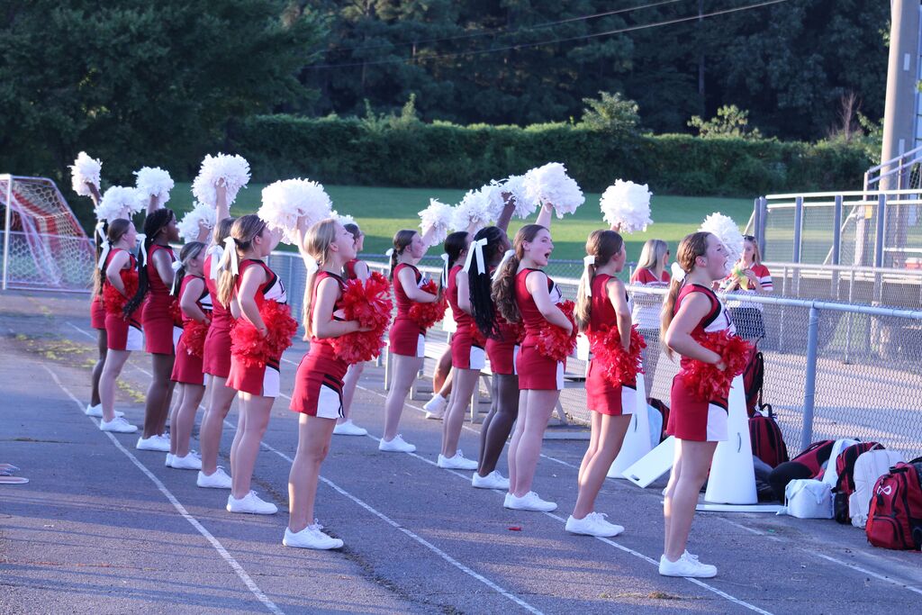 Cheerleaders in red and white uniform lead cheer using pom poms during football game.