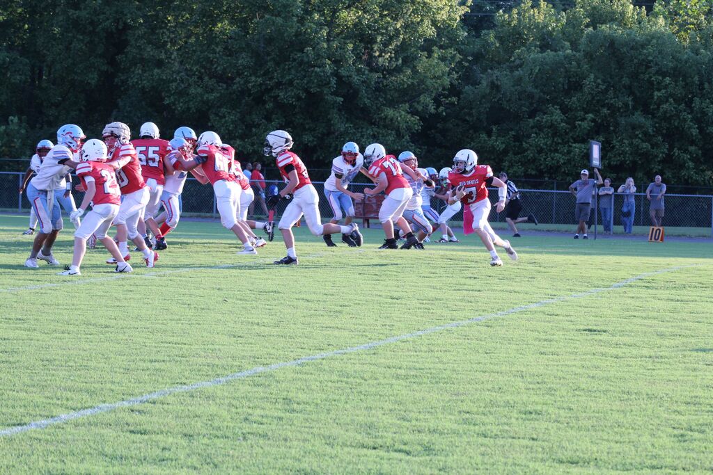 Football player in red and white uniform running the football in a game while the defensive line protects him.