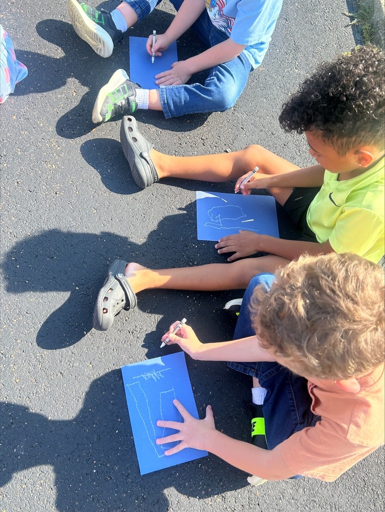Students sit on pavement outdoors, using white markers to draw on blue paper during a hands-on art activity.