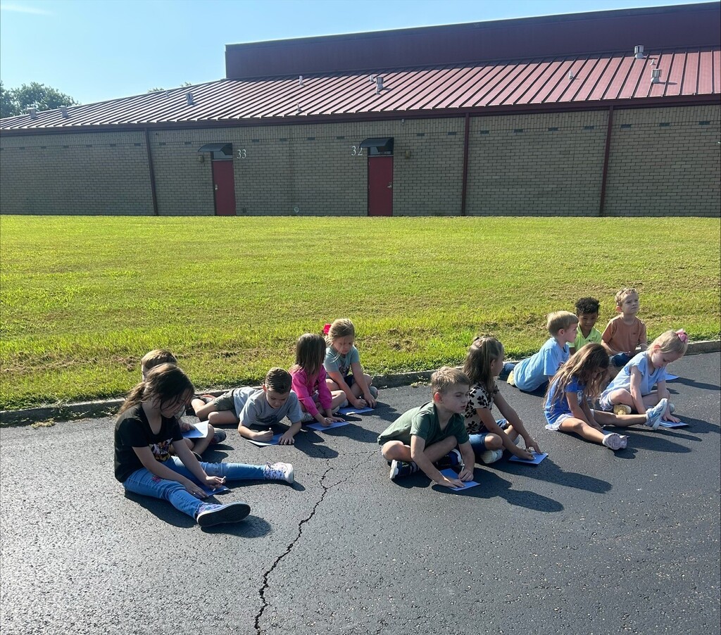 Elementary school students sit on pavement outside a brick school building, writing on blue paper during an outdoor learning activity on a sunny day.