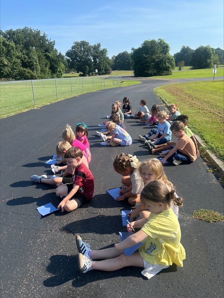 Children sit on an outdoor paved area in the sunshine, drawing with markers on blue paper as part of a group activity.