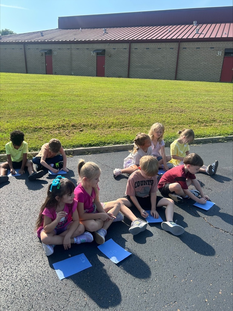 Elementary school students sit on pavement outside a brick school building, writing on blue paper during an outdoor learning activity on a sunny day.