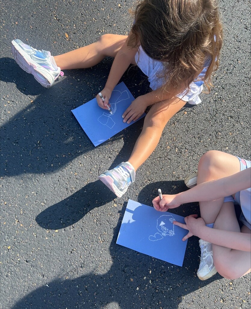 Two young children sit on pavement outdoors, drawing with white crayons on blue paper during a sunny outdoor learning activity.