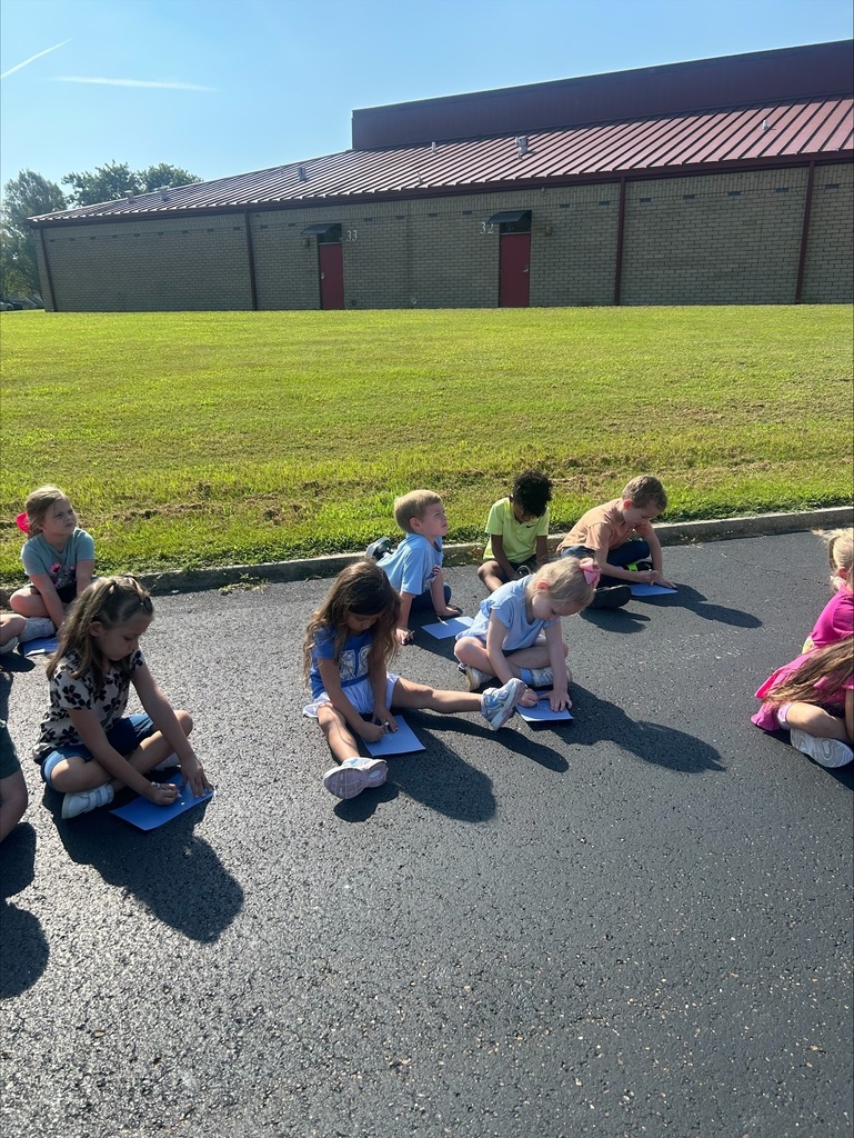 Children sit outside on pavement with blue paper and markers, participating in a hands-on drawing activity.