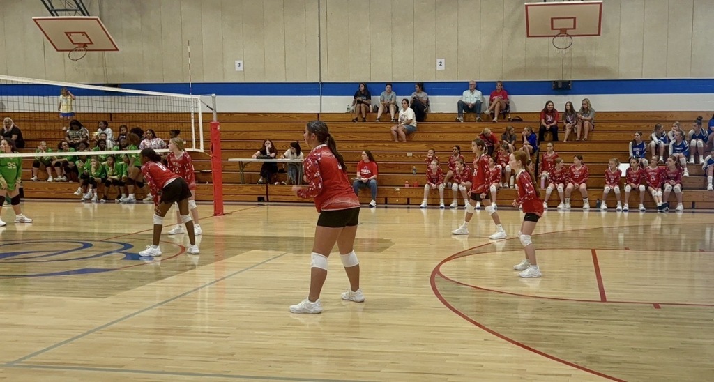 OCMS volleyball players in red uniforms prepare to receive the ball during a game in a school gym, with teammates, opponents, and spectators visible in the background.