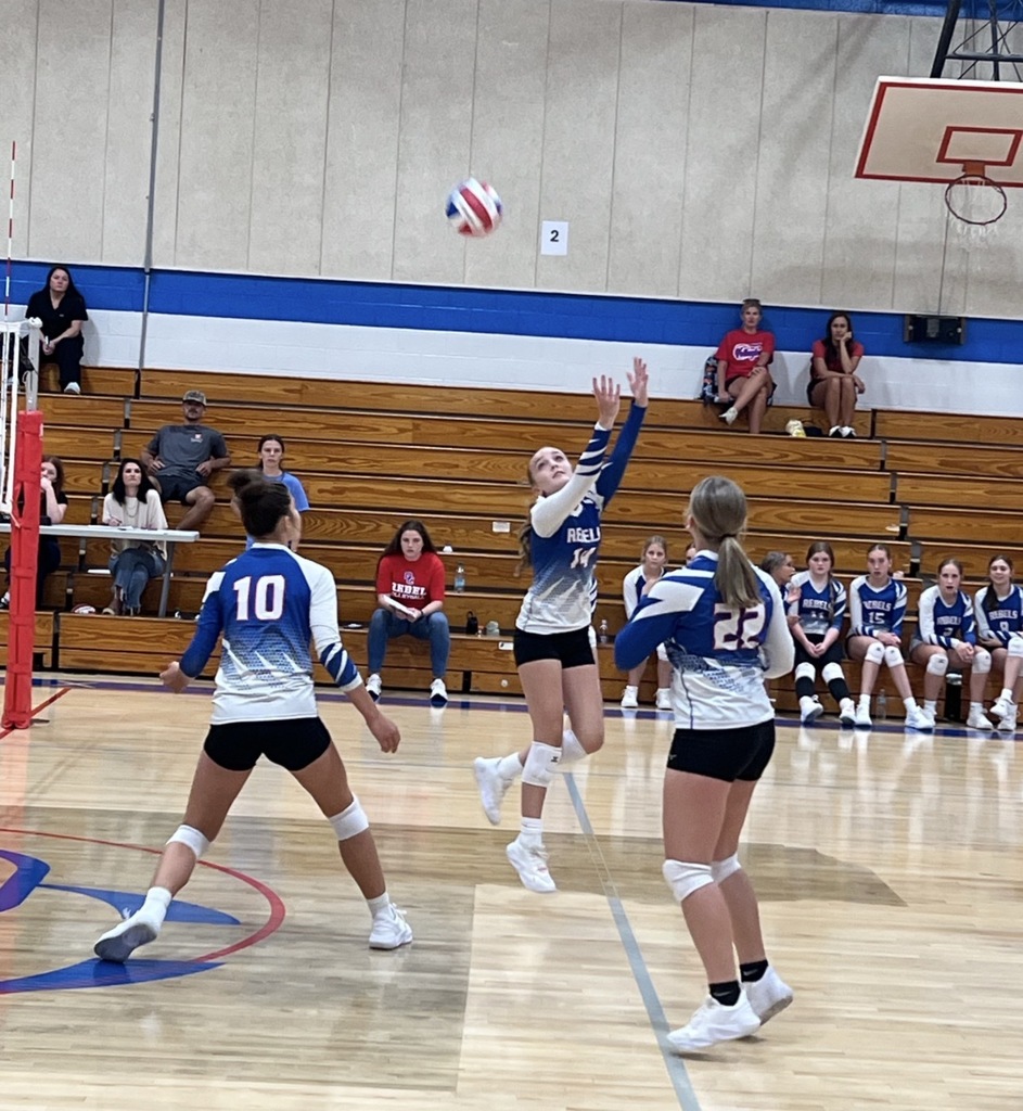 A volleyball player in a blue-and-white Rebels uniform jumps to play the ball as two teammates prepare nearby during a game in a school gym. Teammates and spectators watch from the bleachers.