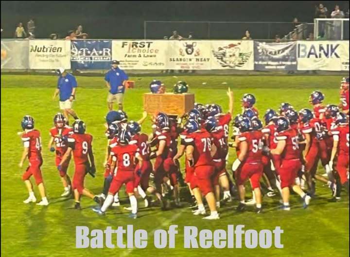 A youth football team in red uniforms gathers on the field during a nighttime game, with coaches and spectators in the background. The image includes the text “Battle of Reelfoot.”