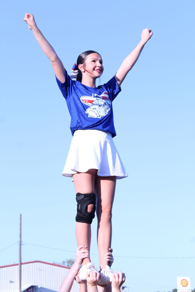 A cheerleader in a blue shirt and white skirt is lifted into the air by teammates, raising both arms in a cheer pose against a bright blue sky.