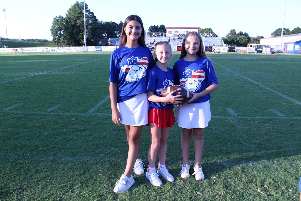 Three young girls stand on a football field wearing matching blue shirts featuring a patriotic bull mascot. The center girl holds a football, while bleachers and a stadium are visible in the background.