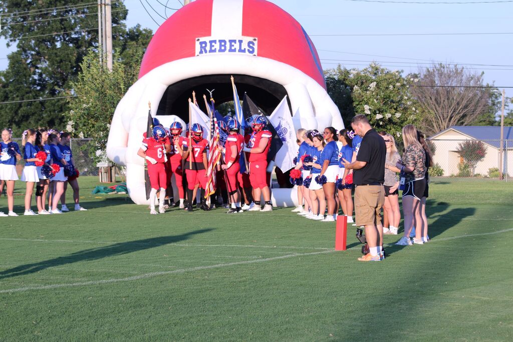 Obion County Central High School football players in red uniforms run onto the field carrying American and team flags, led by cheerleaders holding pom-poms. A large inflatable “Rebels” entrance arch is visible in the background.