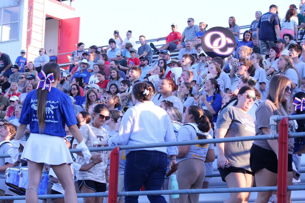 A large crowd of OC supporters fills the bleachers at a sporting event, with cheerleaders and a student pep band playing instruments in the foreground and an OC logo sign displayed above the crowd.