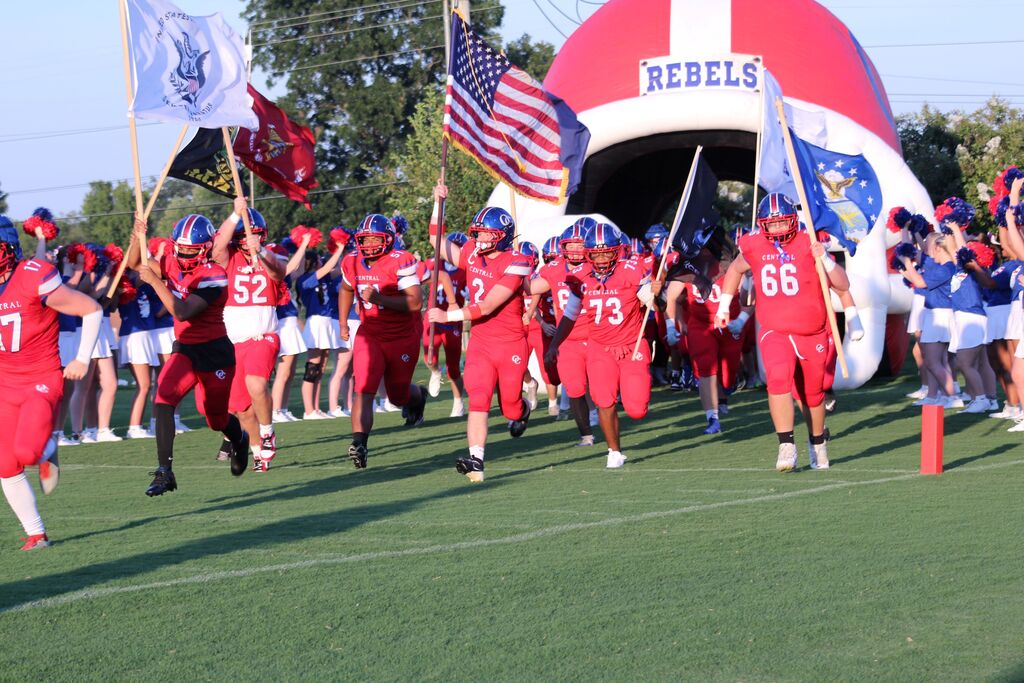 Obion County Central High School football players in red uniforms run onto the field carrying American and team flags, led by cheerleaders holding pom-poms. A large inflatable “Rebels” entrance arch is visible in the background.