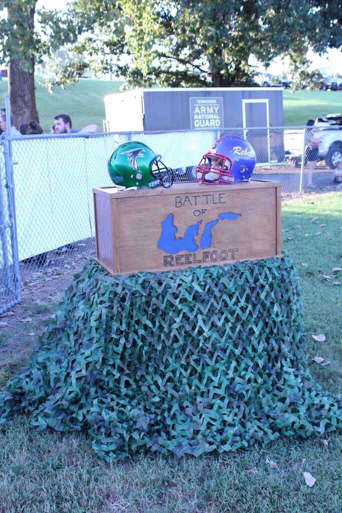 A display for the “Battle of Reelfoot” features green and blue football helmets resting on a wooden box decorated with a map of Reelfoot Lake, surrounded by camouflage netting on a grassy field.