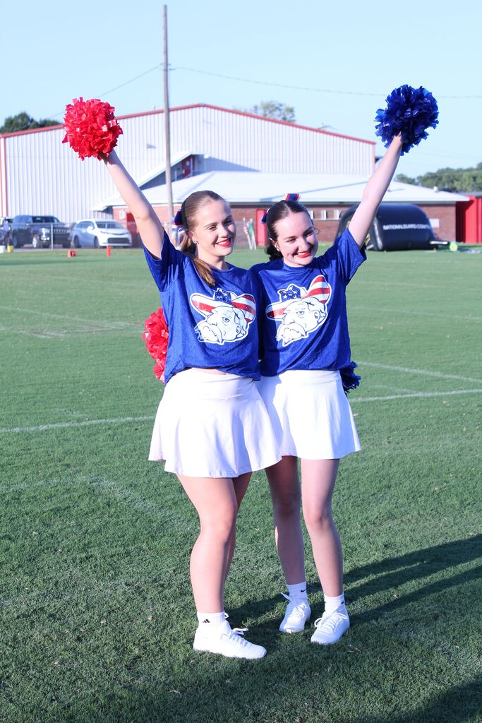 Two cheerleaders in blue shirts and white skirts pose on a football field, smiling and raising red and blue pom-poms. A school building and athletic field are visible in the background.