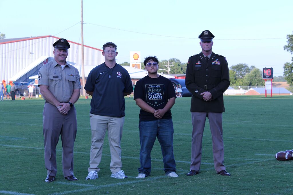 Four people stand on a football field during a ceremony, including two men in military uniforms and two young men wearing athletic and Tennessee Army National Guard shirts. Footballs and a 40-second scoreboard are visible in the background.