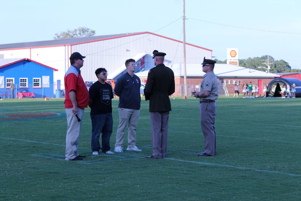 Five people stand together on a football field during a ceremony, including two men in military uniforms and two young men. One young man wears a Tennessee Army National Guard shirt. A school athletic facility, spectators, and football equipment are visible in the background.