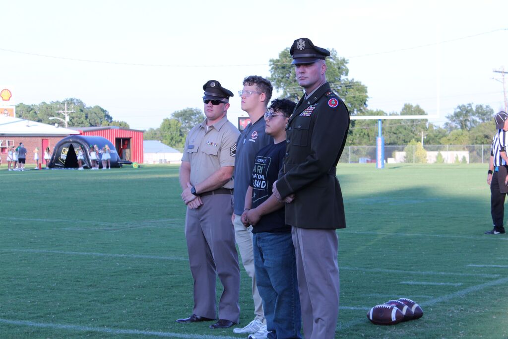 Four people stand at attention on a football field during a ceremony. Two men wear military-style uniforms, while two young men stand between them. Footballs are visible on the field, with a school building, spectators, and athletic equipment in the background.
