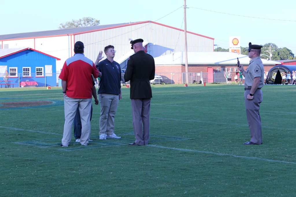 Four people stand on a football field during a ceremony, with two wearing military uniforms. A young man in a school athletic shirt appears to be speaking with the group, while another uniformed man stands nearby holding a phone. Buildings, spectators, and a football field are visible in the background.