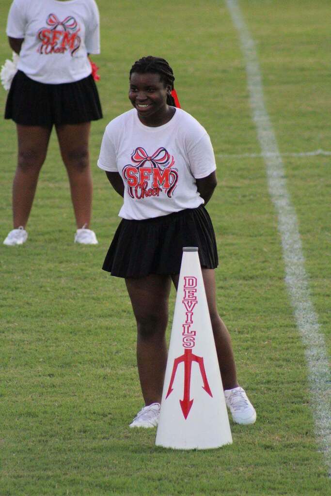 Cheerleader wearing an SFM Cheer shirt and black skirt smiles on the football field beside a white Devils megaphone. Another cheerleader stands in the background.