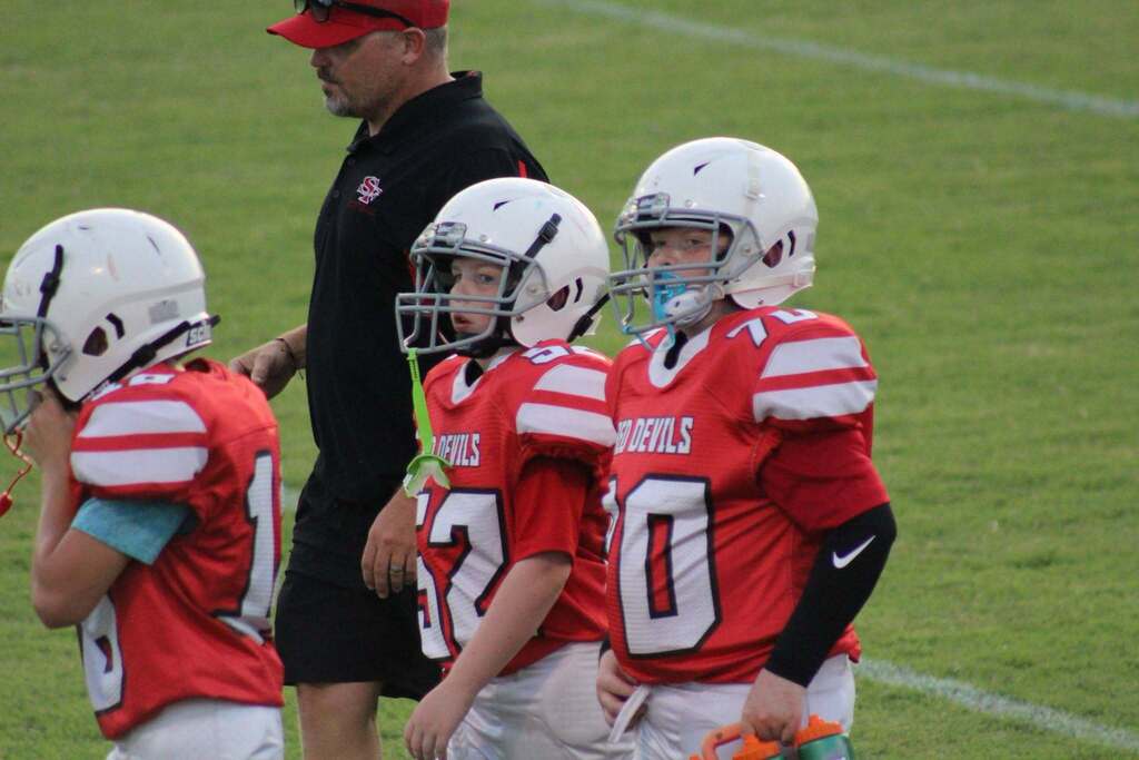 Three youth football players in red Red Devils uniforms and white helmets stand on the sideline with a coach during a game. One player has a blue mouthguard, while the players watch the action on the field.