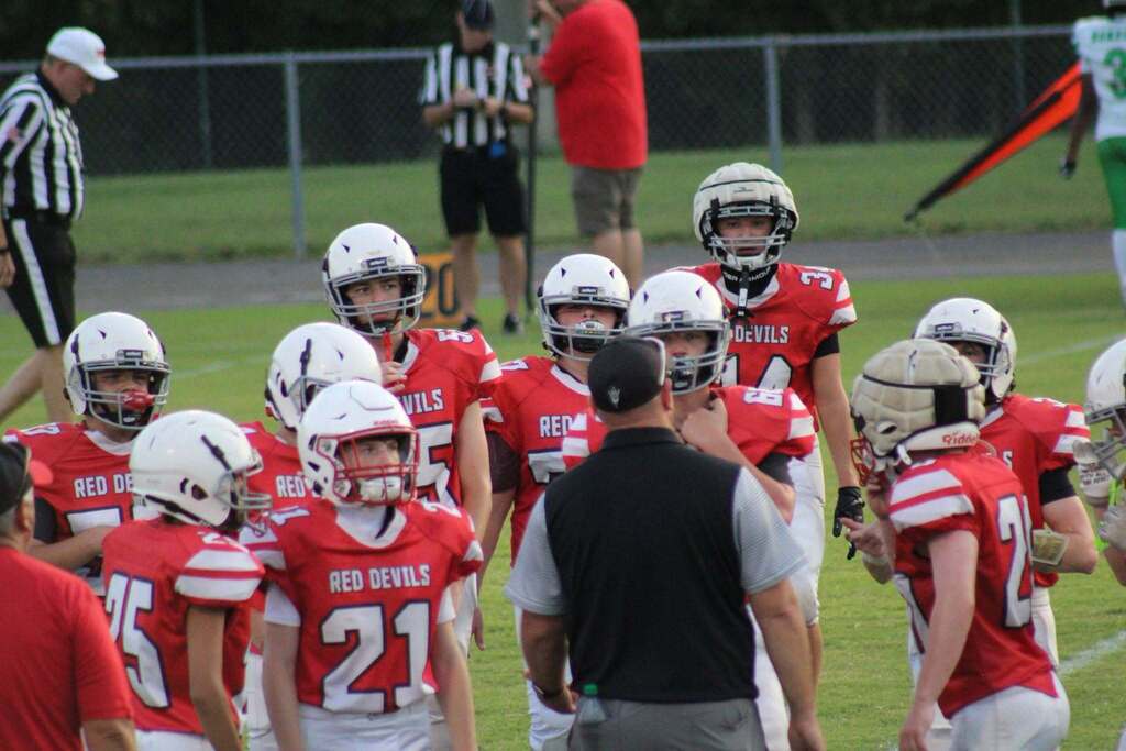 Youth football players in red Red Devils uniforms gather around their coach on the sideline during a game. Players wear white helmets as referees and members of the opposing team are visible in the background.