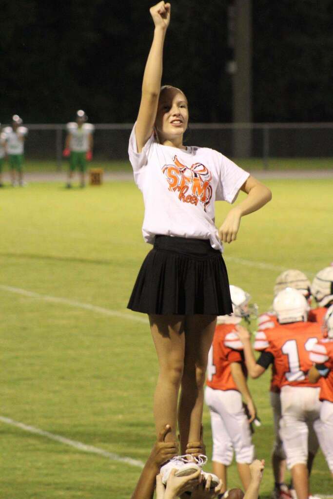 Cheerleader in an SFMS Cheer T-shirt and black skirt raises one fist while being lifted by teammates during a middle school football game. Football players in red uniforms and helmets are visible on the field behind her.