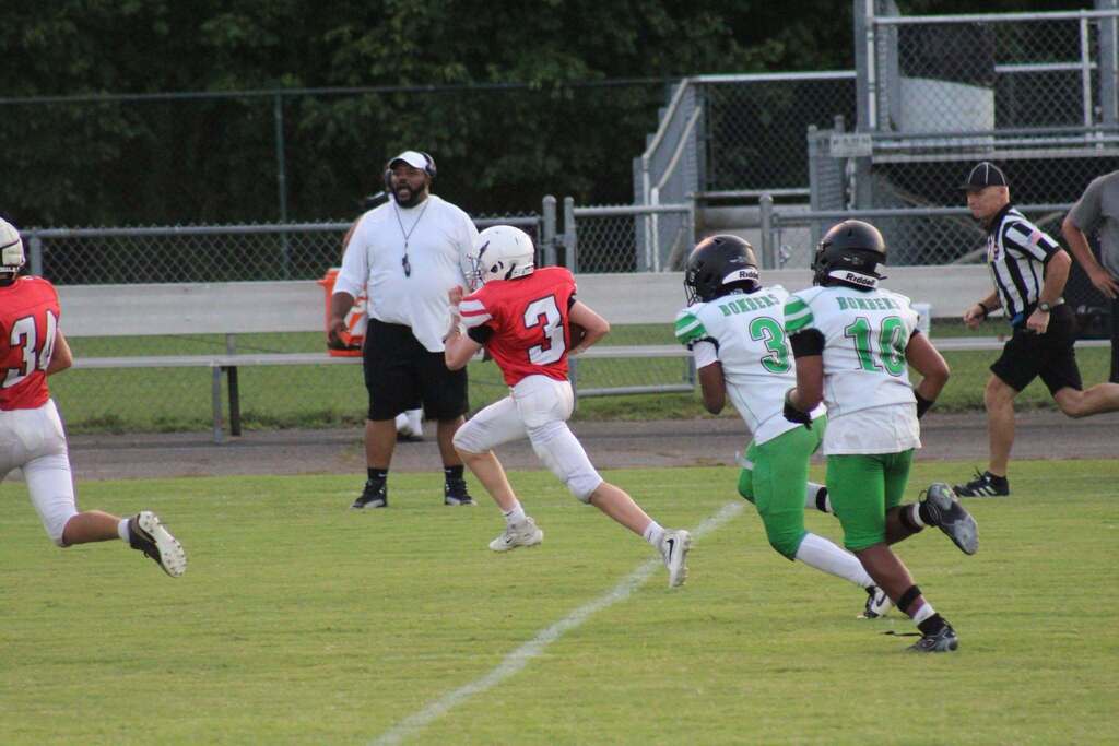 Youth football players in red and green uniforms run across the field during a game. A red-uniformed player wearing number 3 runs with the ball as two green-uniformed defenders pursue him. Coaches and a referee watch from the sideline.