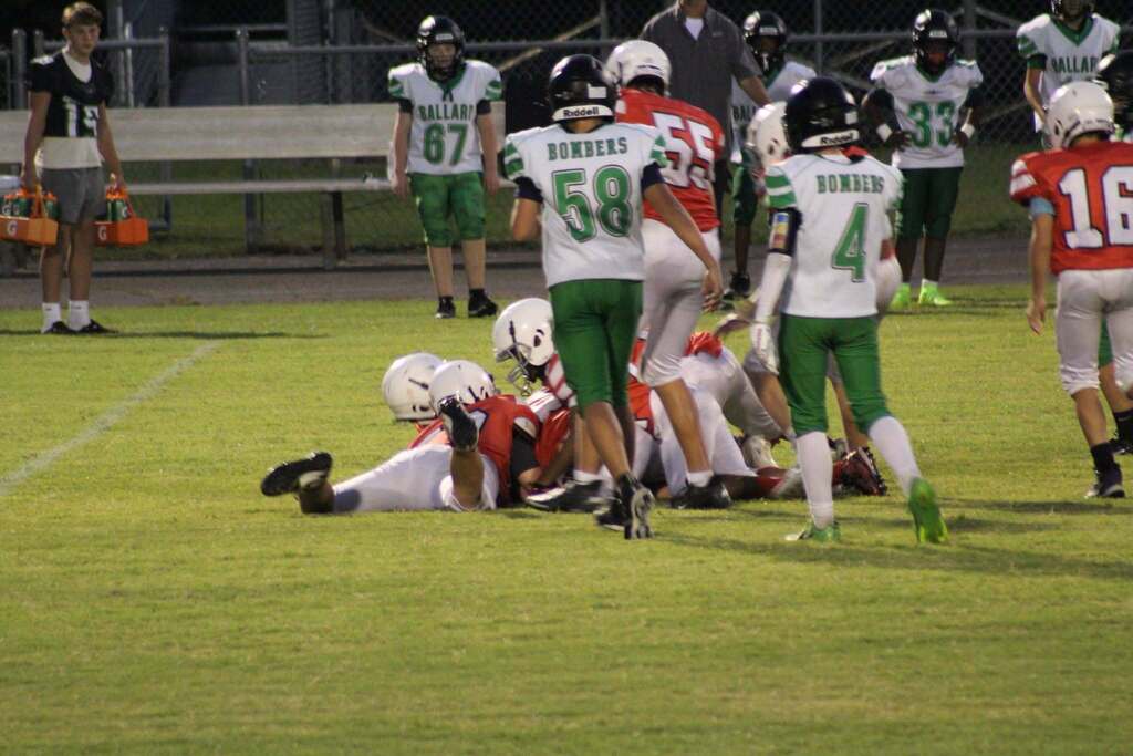 Youth football players in red and green uniforms gather around a tackled player on the field during a game. Ballard Bombers players in white and green uniforms, including numbers 58 and 4, stand near the play while teammates and staff watch from the sideline.