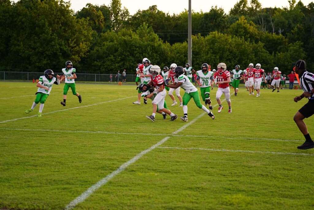 Youth football players in red and green uniforms compete on a grassy field during a game, with several players running toward the play and a referee watching from the right. Trees and spectators are visible in the background.