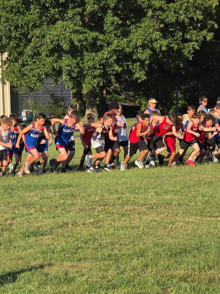 A large group of young cross country runners takes off from the starting line on a grassy field. The runners wear a variety of team uniforms, including blue and red Rebels uniforms, while coaches, parents, and spectators watch from behind. Trees and a school building are visible in the background.
