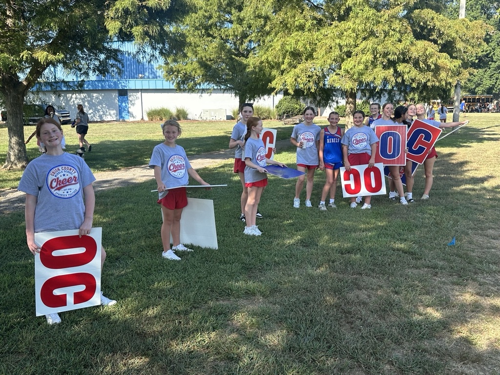 A group of Obion County Middle School cheerleaders stand on a grassy field outside, smiling and holding large red-and-white signs spelling “OC” and “GO” as they cheer on their school. Several students wear matching gray OCMS Cheer shirts and red shorts. Trees provide shade, with a school building and school buses visible in the background.